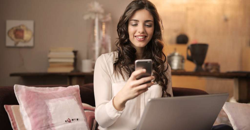 vrouw-zittend-op-de-bank-tijdens-het-kijken-naar-telefoon-met-laptop-op-schoot